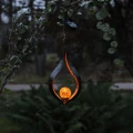 Dekoracyjna lampa solarna LED w formie płomienia z bursztynową kulą, zawieszona wśród zieleni. Delikatne, ciepłe światło podkreśla spokojny, naturalny charakter przestrzeni ogrodowej wieczorem.