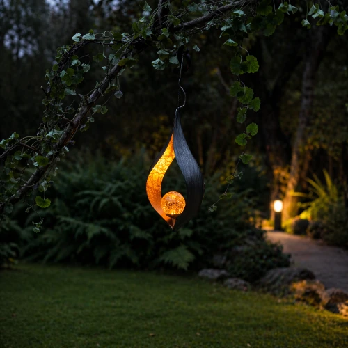 Wisząca lampa solarna LED w formie płomienia z dekoracyjną kulą, zawieszona na gałęzi w ogrodzie. Ciepłe światło tworzy przytulny klimat i subtelnie podkreśla ścieżkę oraz otaczającą zieleń po zmroku.