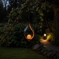 Wisząca lampa solarna LED o smukłej, dekoracyjnej formie płomienia, zawieszona na gałęzi nad ogrodową ścieżką. Ciepłe światło kuli subtelnie oświetla otoczenie, tworząc przytulny klimat po zmroku.