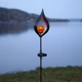 Lampa solarna LED o dekoracyjnej formie płomienia ustawiona na tle spokojnego krajobrazu. Bursztynowa kula emituje ciepłe światło, tworząc nastrojowy akcent w naturalnym otoczeniu.