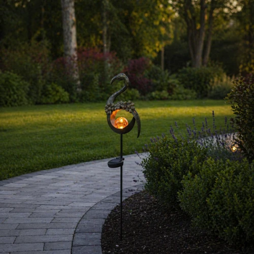 Dekoracyjna lampa solarna LED na szpikulcu w formie stylizowanego ptaka, ustawiona przy ogrodowej ścieżce. Ciepłe światło kuli tworzy przytulny klimat i subtelnie podkreśla roślinność po zmroku.