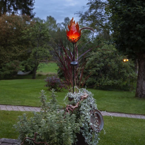 Solarna lampa ogrodowa LED w kształcie płomienia, umieszczona wśród roślin w donicy. Jej ciepłe, bursztynowe światło tworzy przytulny nastrój i subtelnie podkreśla kompozycje roślinne po zmroku.