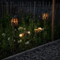 Dwie solarne lampki ogrodowe z metalową osłoną z motywem płomieni, ustawione wśród roślin. Emitują ciepłe światło, które podkreśla naturalny charakter aranżacji po zmroku.