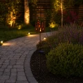 Solarna lampka ogrodowa z metalową osłoną w kształcie płomieni, umieszczona przy ścieżce. Delikatne, ciepłe światło tworzy przytulny klimat w wieczornym ogrodzie.