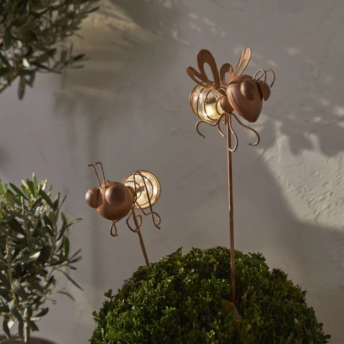 Dwie solarne dekoracje ogrodowe na metalowych prętach w kształcie mrówki i muchy. Umieszczone wśród zieleni, emitują ciepłe światło, które tworzy przyjemny, nastrojowy efekt po zmroku.