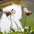 Dwie metalowe dekoracje solarne w formie muchy i mrówki przy oczku wodnym, subtelny akcent do roślin i naturalnych aranżacji ogrodu.