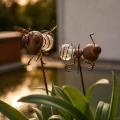 Zbliżenie na solarne dekoracje ogrodowe w kształcie muchy i mrówki. Metalowa konstrukcja z przeszklonym elementem delikatnie odbija światło, tworząc dekoracyjny akcent wśród roślin przy oczku wodnym.