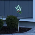 Solarna lampka ogrodowa w kształcie gwiazdy z zielonym światłem, ozdobiona żabką i kwiatami, ustawiona przy elewacji i rabacie, tworzy przyjemny, dekoracyjny akcent i subtelne oświetlenie po zmroku.