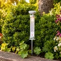 Solarny termometr ogrodowy ze skalą w stopniach Celsjusza i Fahrenheita zamontowany na metalowym pręcie wśród zielonych roślin, do pomiaru temperatury powietrza w ogrodzie.