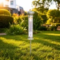 Solarny termometr ogrodowy ze skalą °C i °F na metalowym szpikulcu wbitym w trawnik, ustawiony w nasłonecznionym ogrodzie, do monitorowania temperatury zewnętrznej przy domu i na działce.
