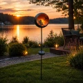 Solarny lampion ogrodowy z dekoracyjnym motywem wróżki w okrągłej oprawie, emitujący ciepłe światło LED, ustawiony na stojaku przy jeziorze w ogrodzie, tworzący nastrojową atmosferę po zmroku.