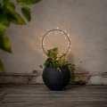 Solarna dekoracja LED w kształcie świetlnego okręgu umieszczona w donicy z zieloną rośliną. Stylowa ozdoba ogrodowa z ciepłym światłem LED tworząca przytulny klimat na tarasie, balkonie lub w ogrodzie.