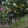 Zbliżenie na solarne dekoracje LED w formie sztucznych ogni z panelem solarnym umieszczone wśród kwiatów i roślin ogrodowych. Delikatne ciepłe światło tworzy nastrojową iluminację i podkreśla wyjątkowy charakter rabaty po zmroku.