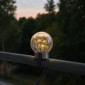 Dekoracyjna lampa solarna LED zamocowana na poręczy balkonu, szklana kula z efektem spękanego szkła emitująca ciepłe światło, nowoczesne oświetlenie na taras i balkon.