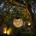 Dekoracyjna lampa solarna LED w formie wiszącej szklanej kuli z ciepłym światłem zawieszona na gałęzi drzewa, nastrojowe oświetlenie ogrodowe na taras, balkon i do strefy relaksu.