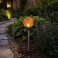 Solarna lampa ogrodowa LED z bursztynową kulą ze spękanego szkła oświetlająca rabatę kwiatową przy kamiennej ścieżce w ogrodzie po zmroku.