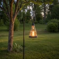 Solarny lampion ogrodowy LED z efektem płomienia zawieszony na metalowej sztycy w ogrodzie, zapewniający ciepłe i dekoracyjne oświetlenie po zmroku.