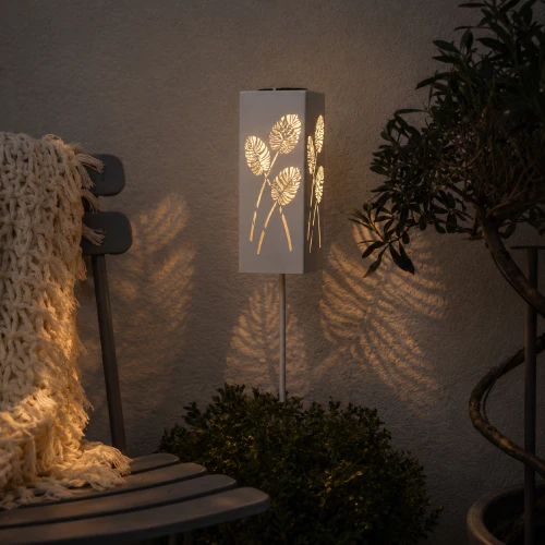 Solarny lampion LED z dekoracyjnym wzorem liści ustawiony w ogrodzie przy tarasie, tworzący efektowną grę świateł i cieni po zmroku.