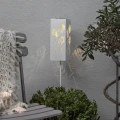 Solarny lampion LED z wycinanym wzorem liści ustawiony przy tarasie, tworzący dekoracyjne refleksy świetlne na ścianie i wśród roślin.