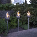 Solarne lampy LED z efektem płomienia ustawione na metalowych sztycach wśród roślin ogrodowych.