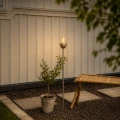 Solarna lampa ogrodowa LED z efektem płomienia ustawiona przy tarasie wśród roślin i dekoracji ogrodowych.