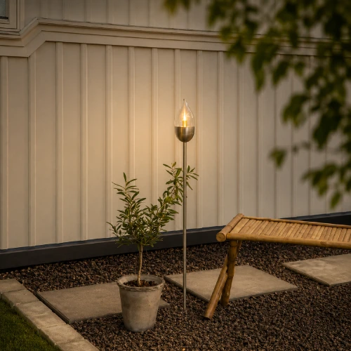 Solarna lampa ogrodowa LED z efektem płomienia ustawiona przy tarasie wśród roślin i dekoracji ogrodowych.