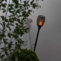Solarna lampa ogrodowa LED z efektem płomienia zamontowana na ścianie przy ozdobnych krzewach w ogrodzie.