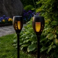 Dwie solarne lampy ogrodowe LED z efektem płomienia ustawione wśród roślin i krzewów w ogrodzie.