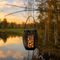 Solarna lampa LED z efektem płomienia zawieszona na gałęzi nad jeziorem o zachodzie słońca.
