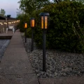 Solarne lampy ogrodowe LED z efektem płomienia ustawione wzdłuż ścieżki przy rabacie i żywopłocie w ogrodzie o zmierzchu.