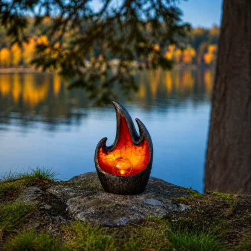 Dekoracyjna lampa solarna LED w formie płomienia ustawiona na kamieniu nad brzegiem jeziora. Ciepłe, bursztynowe światło tworzy spokojny, nastrojowy klimat i subtelnie podkreśla naturalne otoczenie.