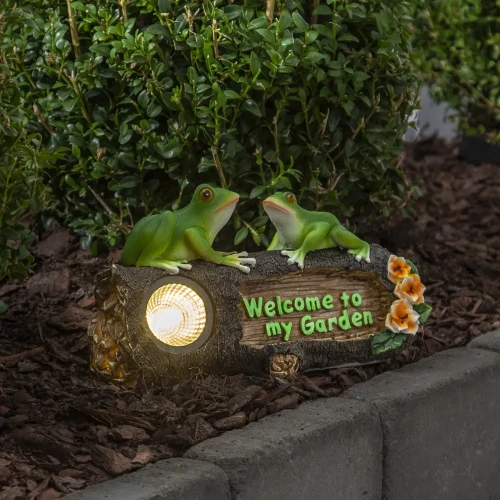 Solarna dekoracja ogrodowa z żabkami i podświetlanym napisem „Welcome to my Garden” ustawiona przy rabacie ogrodowej.