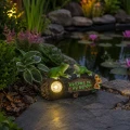 Solarna dekoracja ogrodowa z żabkami i podświetlanym napisem „Welcome to my Garden” ustawiona przy oczku wodnym w ogrodzie.