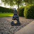 Solarna dekoracja ogrodowa Buddha LED z podświetlaną szklaną kulą ustawiona na kamieniach pod drzewem w ogrodzie.