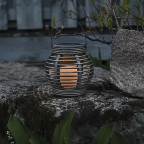 Solarny lampion LED ustawiony na kamiennym murku w ogrodzie, emitujący ciepłe światło po zmroku.