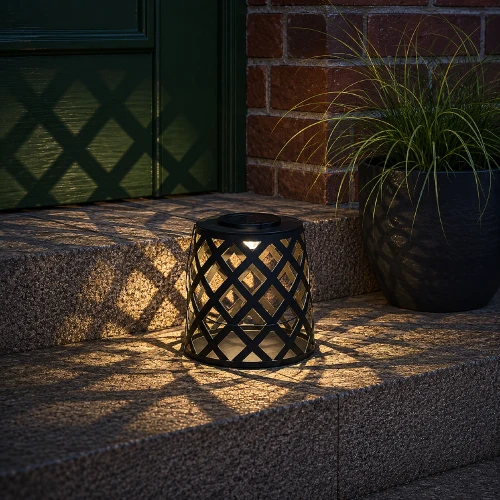 Solarny lampion LED z czarną metalową obudową ustawiony na schodach przed wejściem do domu, emitujący ciepłe światło.