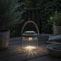 Dekoracyjny lampion solarny LED ze szklaną obudową i jutowym uchwytem ustawiony na stole tarasowym w przytulnej wieczornej aranżacji.