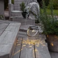 Dekoracyjny lampion solarny LED ze szklaną obudową i metalowym uchwytem ustawiony przy tarasowych meblach, tworzący przytulną atmosferę po zmroku.