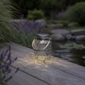 Szklany lampion solarny LED ustawiony na drewnianym pomoście przy oczku wodnym, tworzący nastrojowe oświetlenie ogrodu po zmroku.