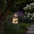 Szklany lampion solarny LED w formie słoika zawieszony na gałęzi drzewa wśród kwitnących roślin ogrodowych po zmroku.