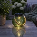 Zielony lampion solarny LED z jutowym uchwytem na drewnianym stole w ogrodzie o zmierzchu.