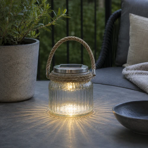 Szklany lampion solarny LED z jutowym uchwytem ustawiony na drewnianym stole tarasowym w otoczeniu roślin doniczkowych po zmroku.