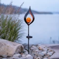Dekoracyjna lampa solarna w kształcie płomienia z podświetloną bursztynową kulą, osadzona wśród jasnych kamieni i traw ozdobnych nad brzegiem zbiornika wodnego.