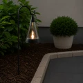 Solarny lampion ogrodowy LED z efektem płomienia zawieszony na metalowej sztycy przy ścieżce ogrodowej, tworzący ciepłe i nastrojowe oświetlenie po zmroku.