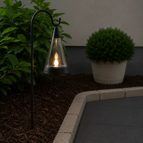 Solarny lampion ogrodowy LED z efektem płomienia zawieszony na metalowej sztycy przy ścieżce ogrodowej, tworzący ciepłe i nastrojowe oświetlenie po zmroku.