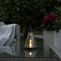 Solarna lampa stołowa z efektem płomienia ustawiona na szklanym blacie tarasu, na tle mebli wypoczynkowych oraz zielonej roślinności ogrodowej o zmierzchu.