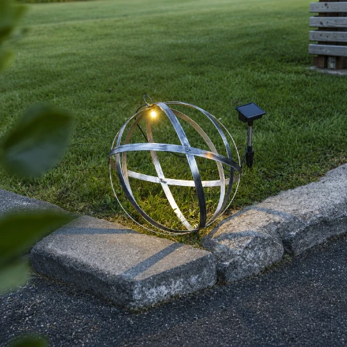 Solarna dekoracja ogrodowa w formie metalowej kuli z podświetleniem LED ustawiona na trawniku przy krawężniku. Delikatne światło wydobywa kształt ażurowej konstrukcji i tworzy subtelne refleksy na metalowych obręczach.