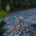 Solarna kula ogrodowa wykonana z przecinających się metalowych obręczy ustawiona przy kamiennym rabacie. Niewielkie źródło światła LED rozświetla konstrukcję i eksponuje jej geometryczny kształt po zmroku.