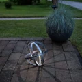 Solarna kula LED z metalowych obręczy ustawiona na kostce brukowej obok donicy z trawą ozdobną. Światło wewnątrz dekoracji podkreśla jej przestrzenną formę i tworzy subtelny wzór cieni na podłożu.