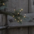 Zbliżenie solarnej dekoracji LED Sztuczne Ognie FIREWORK® umieszczonej na gałązce iglaka w drewnianej donicy. Ciepłobiałe punkty świetlne tworzą subtelny efekt rozświetlonych iskier na tle drewnianej ściany.